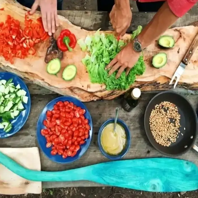Preparing fresh salad ingredients: peppers, lettuce, avocado, tomatoes, cucumbers, dressing and toasted seeds.